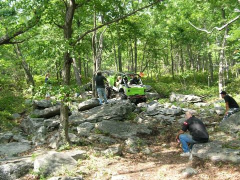 1995 Ford Bronco - A group watches a green off-road vehicle traverse a challenging rocky terrain in a forest.