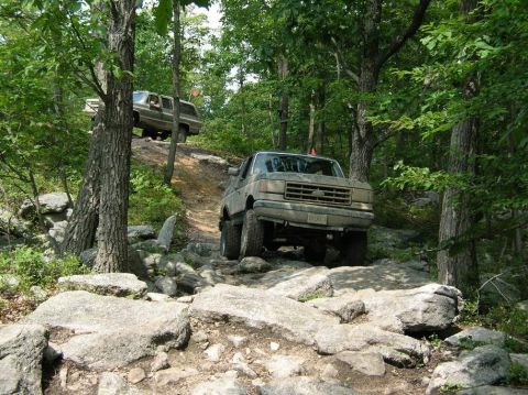 1995 Ford Bronco - A mud-coated full-size Ford Bronco crawls down a rock ledge on a wooded trail, front wheels dropping onto slab granite with the front bumpe…