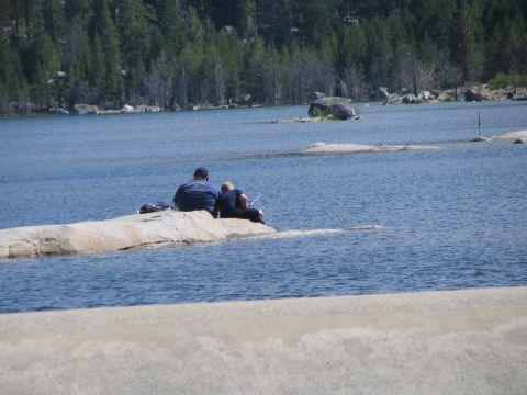 1967 Ford Bronco - Two people enjoy a day of fishing while sitting on a rock in a calm lake, with a forested shoreline in the background.