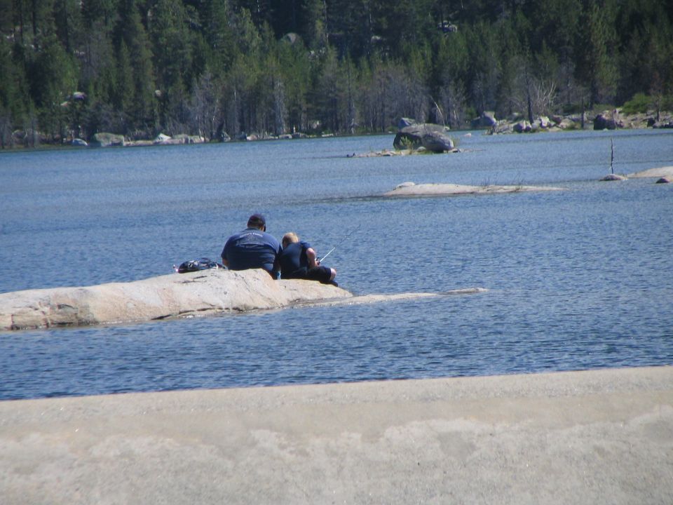 Two people fishing from a granite outcrop on a pine-rimmed mountain lake