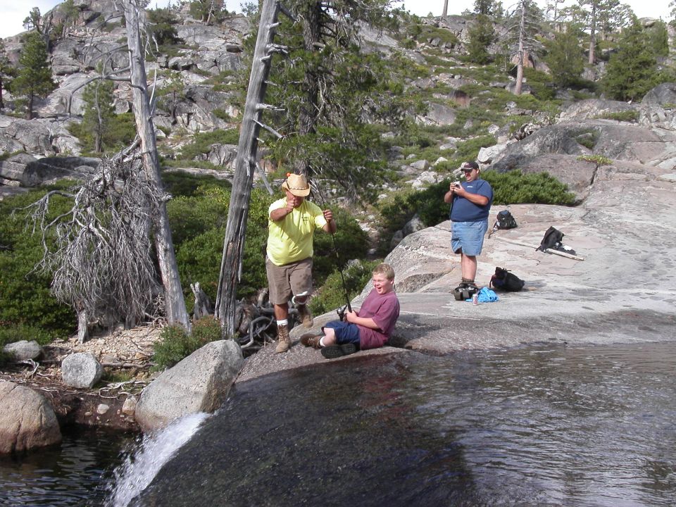 Three people fishing and relaxing on granite rocks beside an alpine pool and waterfall