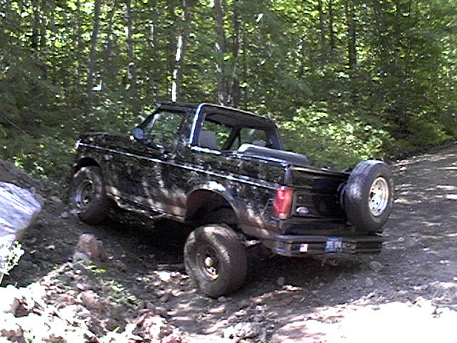 Black Ford Bronco with a spare tire mounted on the rear, driving on a dirt trail in a wooded area