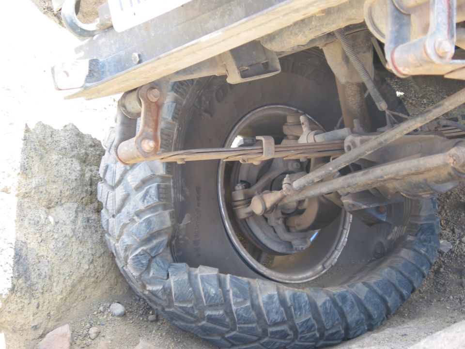 Underside view of leaf-sprung solid front axle, steering linkage and mud-terrain tire on black steel wheel