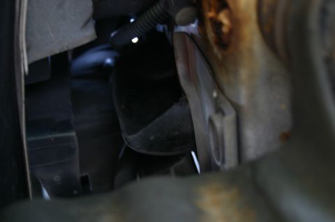 1994 Ford Bronco - A dark, close-up view into a tight engine bay or undercarriage area showing black plastic components, a ribbed conduit hose, and a rusty me…