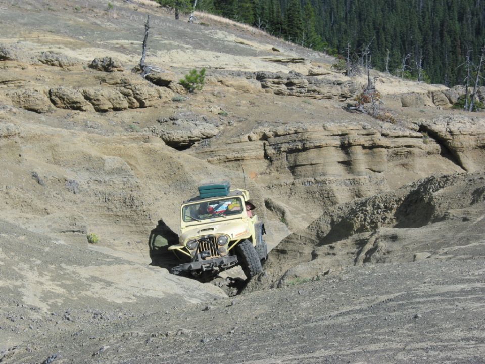 Pale yellow open Jeep CJ dropping off an eroded rock ledge on a forested mountain trail