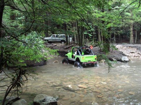 1995 Ford Bronco - A lime-green topless Jeep CJ/Wrangler with a roll bar, orange whip flag and tall mud tires fords a rocky creek in a shaded hardwood forest,…