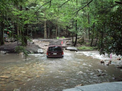 1995 Ford Bronco - Jeep Cherokee XJ navigates a rocky stream bed surrounded by dense woods.