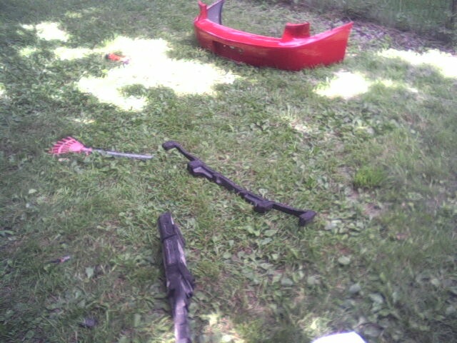 Red car bumper and yard tools on grass. Includes a rake and a black metal bar