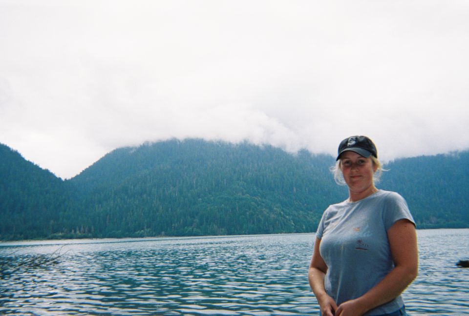 Woman in gray shirt and dark cap standing by a calm lake with fog-shrouded forested mountains