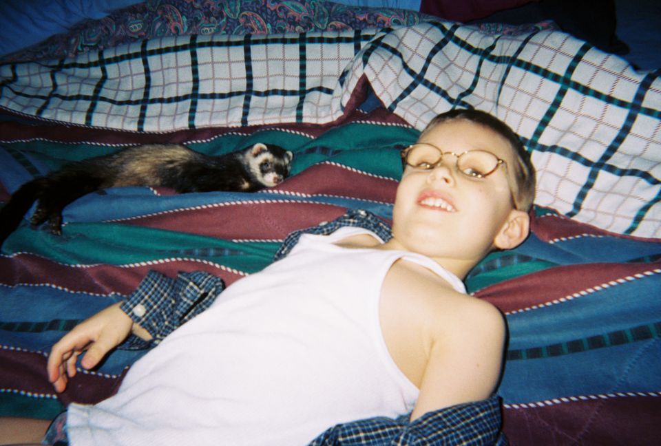 Child with glasses and a ferret lie on a bed with striped blankets