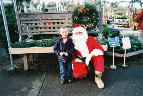 1990 Ford Bronco - A smiling boy in a navy jacket and jeans stands beside a kneeling Santa Claus in a red suit at an outdoor garden center, in front of a wood…