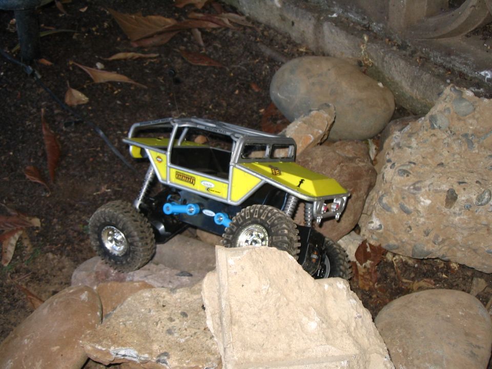 Yellow-bodied RC crawler with chrome cage and blue links scaling rocks among leaves