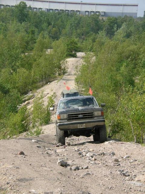 1995 Ford Bronco - Two trucks navigate a rocky, off-road trail with trees and a building in the background.