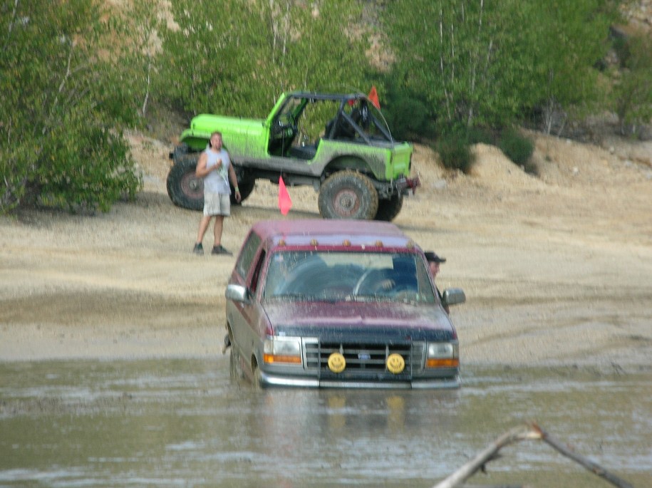 Maroon 1995 Ford Bronco stuck in mud, front view. Green Jeep in background