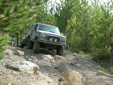 1995 Ford Bronco - This modified SUV navigates a challenging, rocky off-road path surrounded by dense forest.