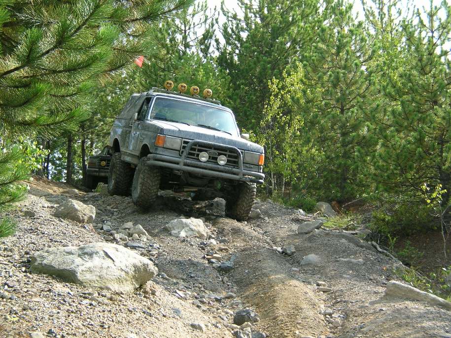 off-road vehicle with roof lights driving on a rocky trail with trees around. another vehicle is behind it