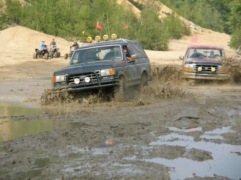 1995 Ford Bronco - Two off-road vehicles navigate a muddy track with ATV riders in the background.