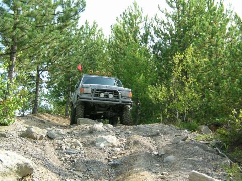 1995 Ford Bronco - A gray Ford Bronco navigates a challenging rocky terrain surrounded by pine trees.