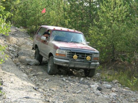 1995 Ford Bronco - Three-quarter front view of a dust-covered maroon full-size Ford Bronco picking its way down a loose rocky bank, with the driver leaning ou…