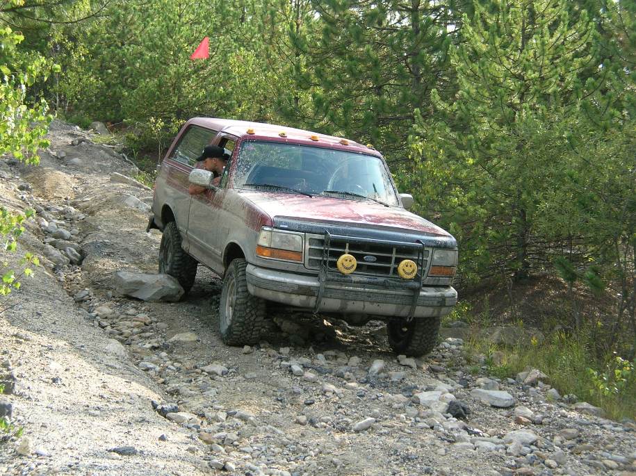 Maroon Ford Bronco with smiley-face light covers descending a rocky bank, driver leaning out