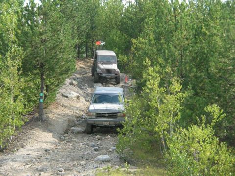 1995 Ford Bronco - A silver pickup truck navigates a rocky off-road trail, with a Jeep following closely behind through the trees.