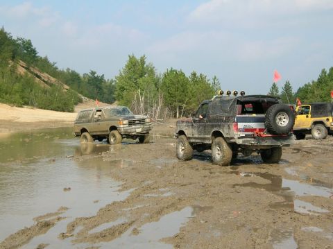 1995 Ford Bronco - Several off-road vehicles navigate a challenging muddy terrain with trees and a sandy embankment in the background.