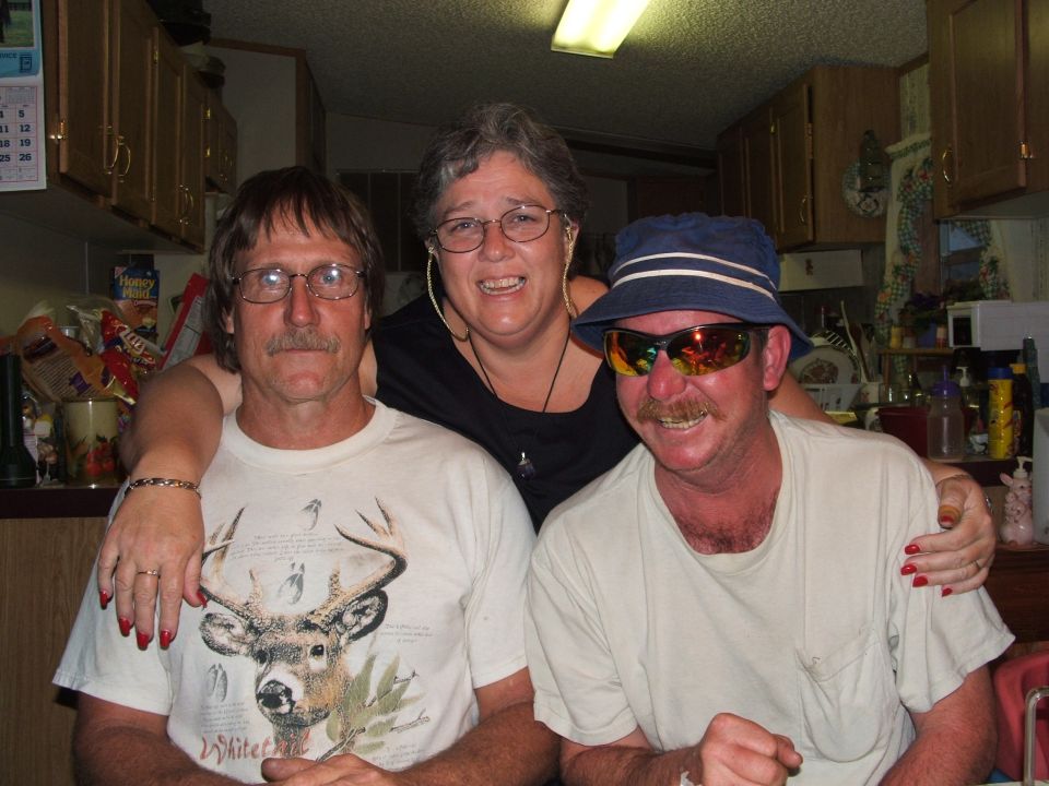 Three people pose for a photo indoors. One man wears a deer t-shirt, another wears sunglasses and a hat