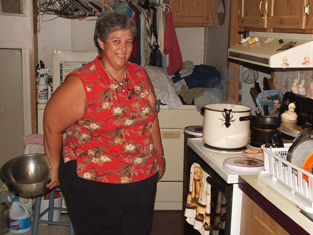 Woman in red floral shirt and black pants standing in a cluttered kitchen with laundry and appliances