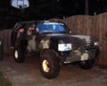 1995 Ford Bronco - Very small, low-resolution night photo of a lifted dark full-size SUV parked on grass beside a wooden fence, its headlights and a roof-moun…