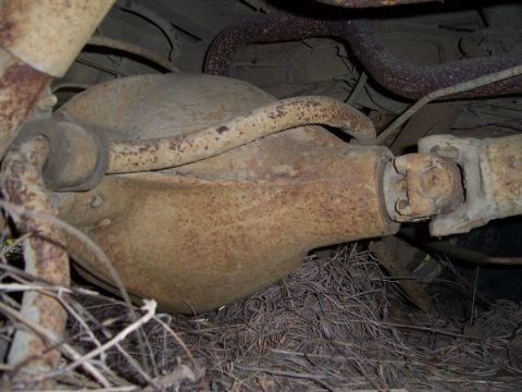 1983 Ford Bronco - Underbody close-up of a rusty, dirt-caked differential pumpkin on a solid axle, with the driveshaft U-joint and yoke at the pinion on the r…