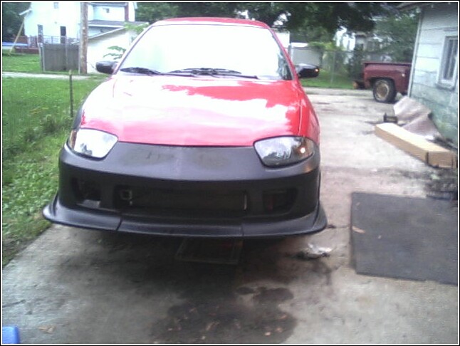 Red car with aftermarket black front bumper and lip kit, parked on concrete