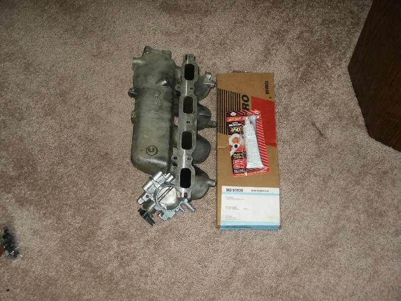 Aluminum intake manifold with throttle body beside a Fel-Pro gasket box and RTV sealant on carpet.