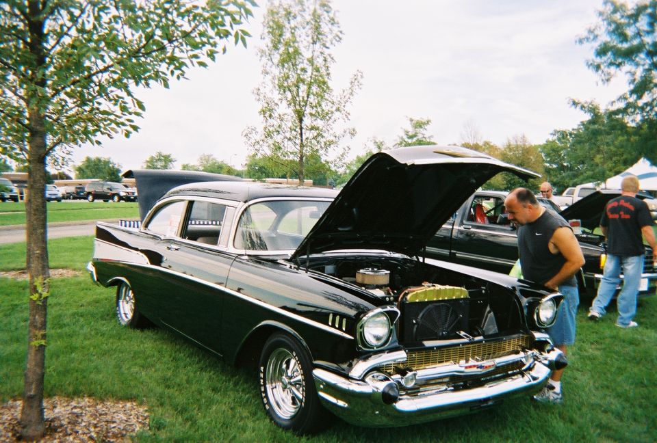 Black 1957 Chevrolet Bel Air with hood open, parked on grass at a car show. Another car and people are visible in the background