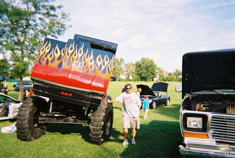 Monster truck with flame decals, lifted with large tires, parked on grass. A man stands next to it