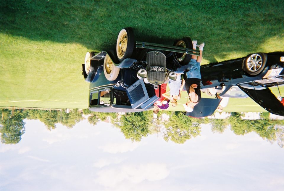Dark blue vintage car with whitewall tires and a license plate reading 239-304. People are gathered around it, and a laptop is on the hood. A modern black car is partially visible to the right