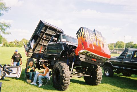 1996 Ford F-150 - A heavily modified dump truck with flame decals on its raised hood and oversized tires is displayed at an outdoor event.