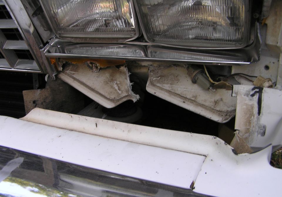 Open front clip of a vehicle showing dual headlights, torn fender metal and detached white trim