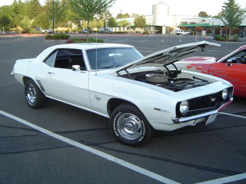 1964 Ford Fairlane - A white 1969 Chevrolet Camaro SS is parked with its hood open, revealing the engine, next to another car with flame decals.