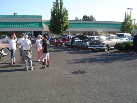 1964 Ford Fairlane - A group of people stand around various vintage automobiles displayed in a parking lot on a sunny day.