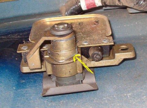 1988 Ford Bronco - Close-up of a gold-cadmium-plated door latch or hood latch mechanism sitting on a blue painted panel, with a yellow drawn arrow pointing at…