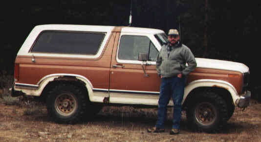 Man in cap and jacket leans against a brown and white Ford Bronco. The vehicle has a removable hardtop