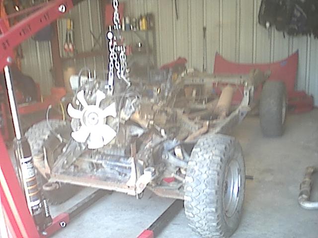 Bare vehicle chassis with engine fan visible in garage, red engine hoist and chains overhead