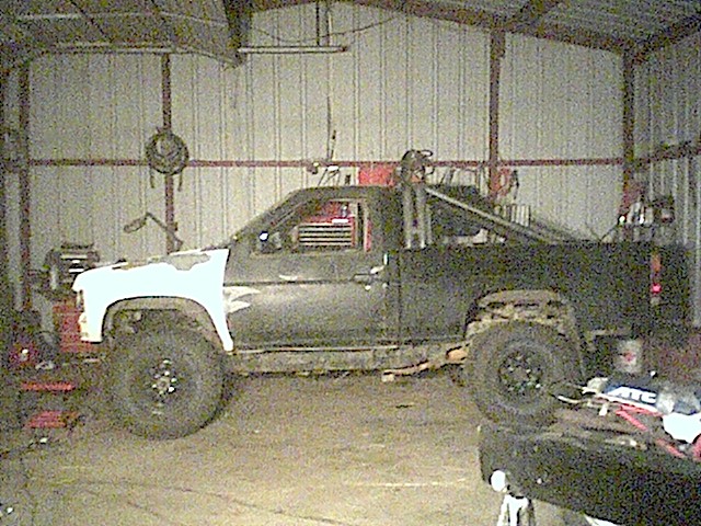 Side view of a dark pickup with white front fender and mud tires inside a metal garage
