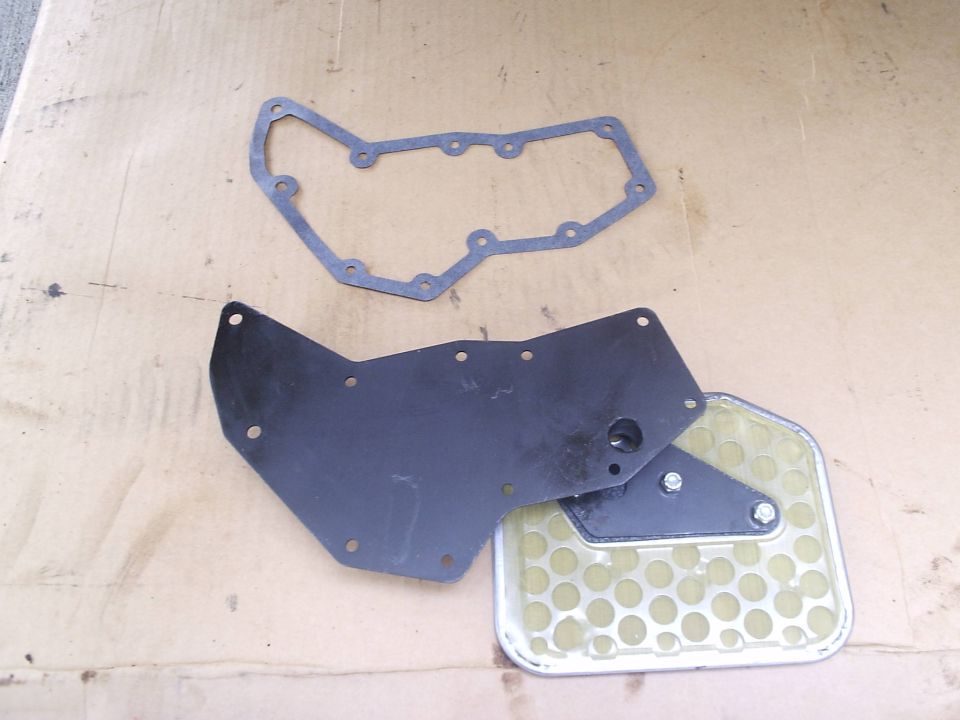 Transmission pan gasket, black metal plate, and new filter with tan screen laid out on cardboard