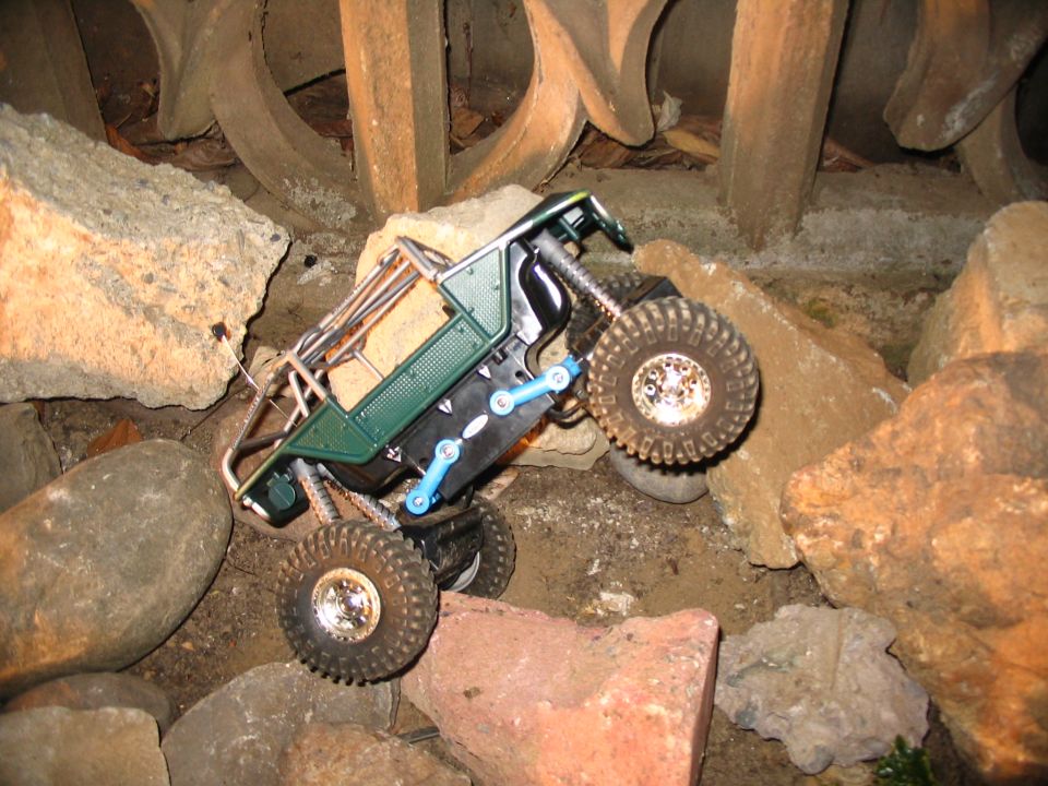 Green RC crawler with blue links climbing nearly vertical over rocks, one front wheel lifted