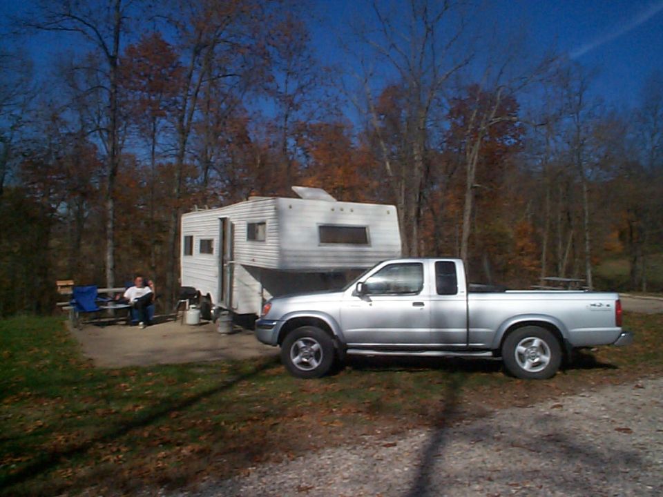 Silver Nissan Frontier extended cab pickup parked at a campsite beside a white fifth-wheel trailer