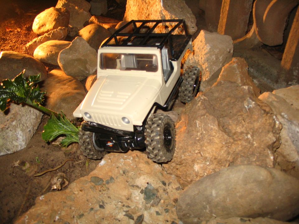 Tan Jeep-style RC crawler body with black roof rack climbing over a large rock at night