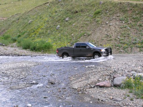 1986 Ford Bronco - A dark gray pickup truck navigates through a rocky stream, likely a Ford F-150 from around 2006.