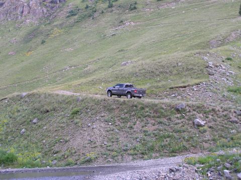 1986 Ford Bronco - A pickup truck drives along a winding dirt road on a steep, grassy mountainside.