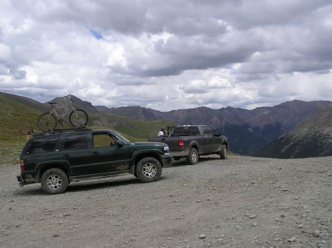 1986 Ford Bronco - Two vehicles are parked on a gravel road in a mountainous area under a cloudy sky.